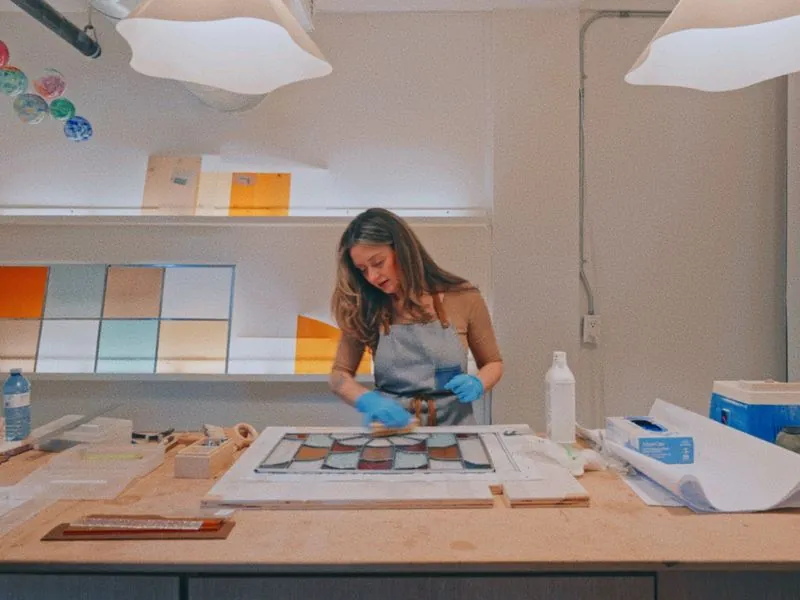 Cleaning a stained glass panel at the workbench, from Art and Motherhood by Studio Propel